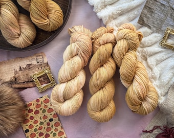 May include: Five skeins of hand-dyed yarn in shades of golden yellow, honey, and soft peach. Two skeins are arranged on a dark circular tray, while three skeins are positioned together in the center, showcasing a twisted, multi-ply fiber texture.
