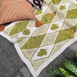 May include: A green and white crocheted throw blanket with a geometric pattern and tassels. The blanket is lying on a gray tiled floor.
