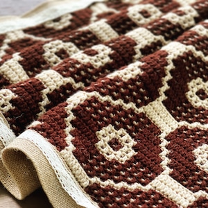 以下が含まれることがあります： 幾何学模様のブラウンとクリーム色のクロシェのテーブルランナー。ランナーはジュートの裏地と白いレースのトリムが付いています。