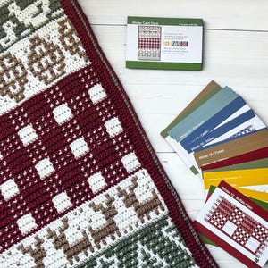 May include: A crocheted blanket featuring a repeating pattern of reindeer, geometric shapes, and buffalo plaid in shades of burgundy, white, olive green, and brown, accompanied by a Winter Card Deck box and a set of instructional pattern cards.