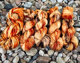May include: Close-up of six skeins of hand-dyed yarn in shades of orange, brown, and cream. The yarn is arranged on a bed of gray and brown stones. The yarn appears soft and textured, ideal for knitting or crochet projects.