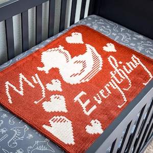 以下が含まれることがあります： オレンジと白の毛糸で編まれたベビーブランケット。"My Everything"という文字と、白の毛糸で編まれたアヒルのデザインが施されています。