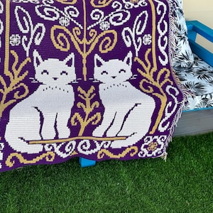 Könnte beinhalten: Eine violett-weiß gewebte Decke mit zwei weißen Katzenfiguren. Die Decke hat ein sich wiederholendes Muster aus goldenen und weißen floralen und geometrischen Designs. Die Decke ist über ein Sofa mit blauem Rahmen und einem Kissen mit einem schwarz-weißen Blättermuster drapiert.