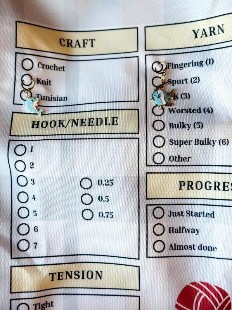 Pode incluir: Uma lista de verifica&ccedil;&atilde;o branca com um cabe&ccedil;alho amarelo para projetos de artesanato. A lista de verifica&ccedil;&atilde;o inclui se&ccedil;&otilde;es para artesanato, fio, gancho/agulha, tens&atilde;o e progresso. A lista de verifica&ccedil;&atilde;o tem um amuleto de golfinho e um amuleto de peixe.