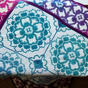 May include: A crocheted blanket with a white background and a repeating pattern of teal and blue flowers. The blanket is made up of squares with different designs.