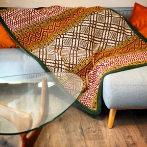 May include: A woven throw blanket with a geometric pattern in shades of red, brown, yellow, and green. The blanket is draped over a grey sofa with orange throw cushions.