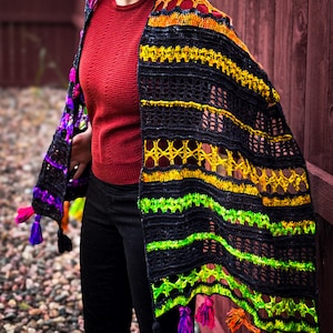 Peut inclure: Un châle crocheté à la main, aux couleurs vives, avec une base noire et des rayures jaunes, vertes et oranges. Le châle présente des pompons décoratifs de différentes couleurs et des motifs ajourés complexes. Le châle est drapé sur les épaules.