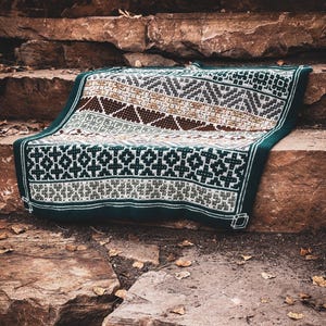 May include: A knitted blanket with a geometric pattern in shades of brown, grey, white, and green. The blanket is folded over a stone surface, showcasing its intricate design and textured appearance. The edges are finished with a dark green border.