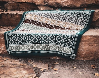 May include: A knitted blanket with a geometric pattern in shades of brown, gray, white, and green. The blanket is folded over a stone surface, showcasing its intricate design and textured appearance. The edges are finished with a dark green border.