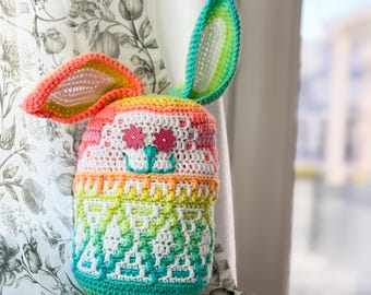 May include: A crocheted rainbow bunny toy with white and pink flower accents. The bunny is sitting on a floral patterned fabric.
