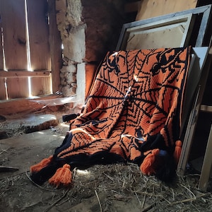 Puede incluir: Una manta de crochet con un patrón geométrico en naranja y negro. La manta está sobre un suelo de madera en un entorno rústico.