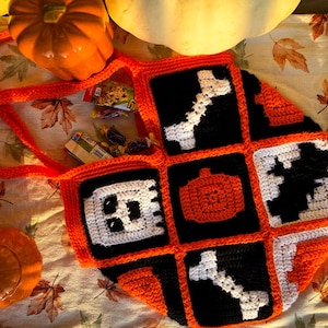 Może przedstawiać: Worek na ramię na Halloween wykonany na szydełku z pomarańczowymi, czarnymi i białymi kwadratami przedstawiającymi ducha, dynię, kości i nietoperza. Worek leży na powierzchni z tkaniny z jesiennymi liśćmi.