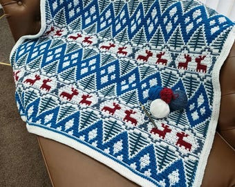 May include: A blue, white, and red crocheted blanket with a geometric pattern of trees and reindeer. The blanket is folded over a brown leather couch. A crochet hook and yarn are visible.