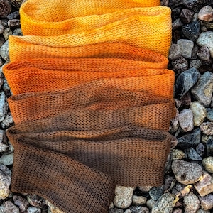 Puede incluir: Una tira larga de tela tejida rectangular que presenta una transición de color degradada que cambia de un amarillo brillante en la parte superior, pasando por tonos naranja, hasta un marrón oscuro profundo en el extremo inferior.