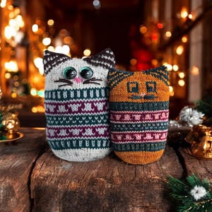 May include: Two knitted cat-shaped ornaments. One is white with a black and brown patterned head and a teal, purple, and white patterned body. The other is orange with a teal and purple patterned body. Both have large eyes.