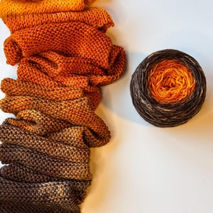 May include: A knitted scarf featuring a color gradient that transitions from bright orange at the top to dark brown at the bottom, positioned next to a round ball of yarn that displays the same color transition from orange to dark brown.