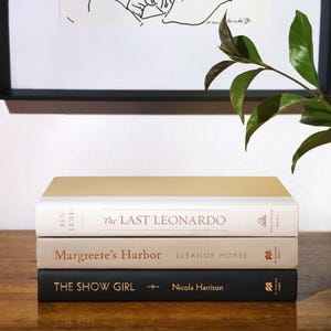 May include: A stack of three hardcover books with neutral-toned covers. The top book has a gold cover, the middle book is beige, and the bottom book is black. Titles include "The Last Leonardo," "Margreete's Harbor," and "The Show Girl."