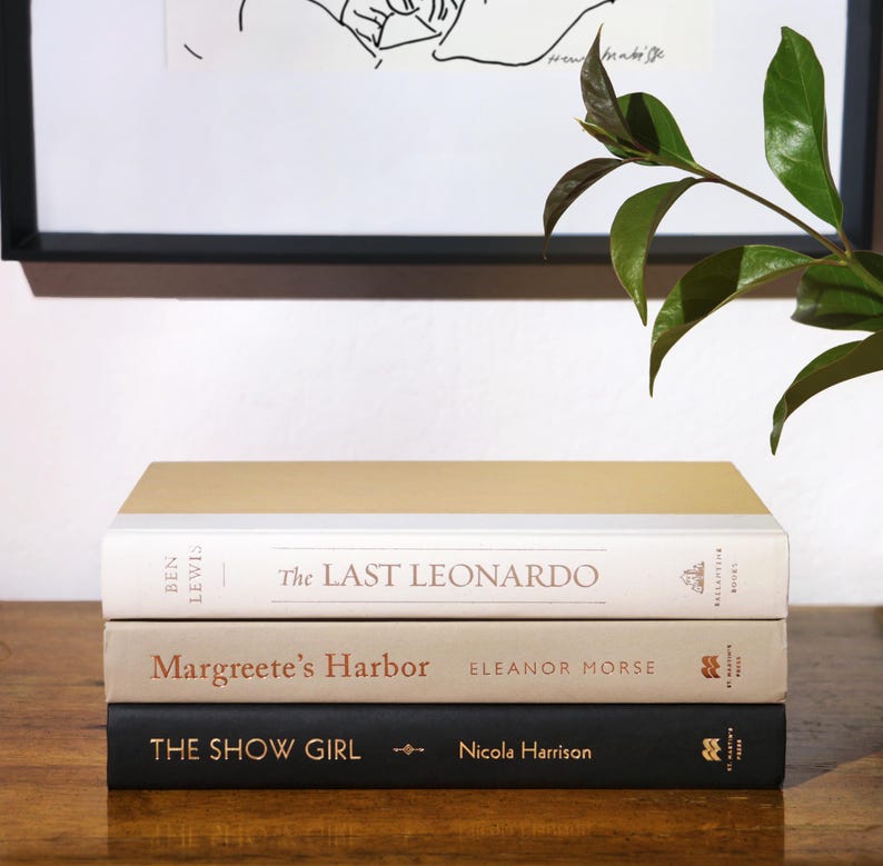 May include: A stack of three hardcover books with neutral-toned covers. The top book has a gold cover, the middle book is beige, and the bottom book is black. Titles include "The Last Leonardo," "Margreete's Harbor," and "The Show Girl."
