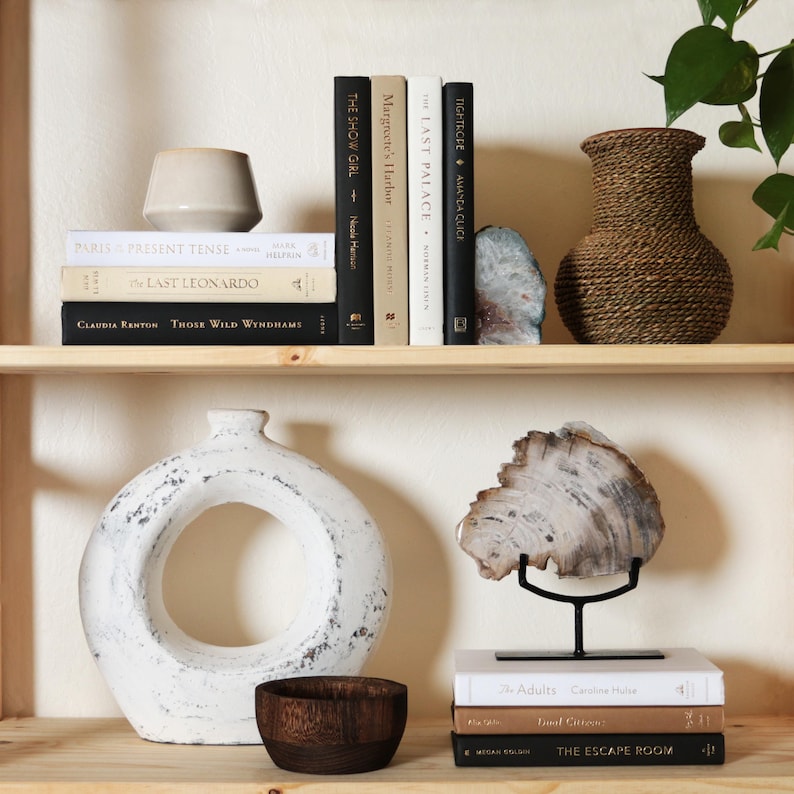 May include: A display of decorative items on wooden shelves. Includes books, a beige vase, a woven vase, a white ceramic vase with a circular opening, a wooden bowl, and a piece of petrified wood on a black stand.