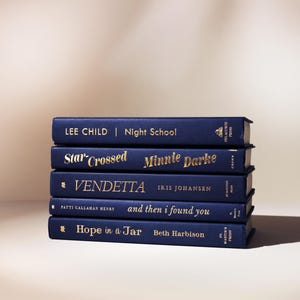 May include: A stack of five hardcover books with navy blue covers and gold lettering. The titles and authors are visible, including "Night School" by Lee Child and "Star-Crossed" by Minnie Darke. The books are stacked neatly on a light-colored surface.