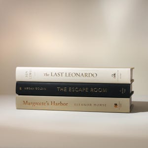 May include: Three hardcover books stacked on top of each other. The top book is white with gold lettering that reads "The Last Leonardo." The middle book is black with gold lettering that reads "The Escape Room." The bottom book is tan with gold lettering that reads "Margreete's Harbor."