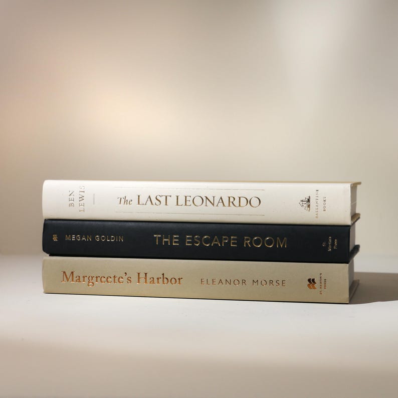 May include: Three hardcover books stacked on top of each other. The top book is white with gold lettering that reads "The Last Leonardo." The middle book is black with gold lettering that reads "The Escape Room." The bottom book is tan with gold lettering that reads "Margreete's Harbor."