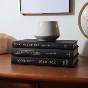 May include: A stack of three black hardcover books with gold lettering, including "What Was Before," "The Da Vinci Code," and "Anne Rice Merrick." A small, light-colored ceramic vase sits atop the books on a wooden surface.