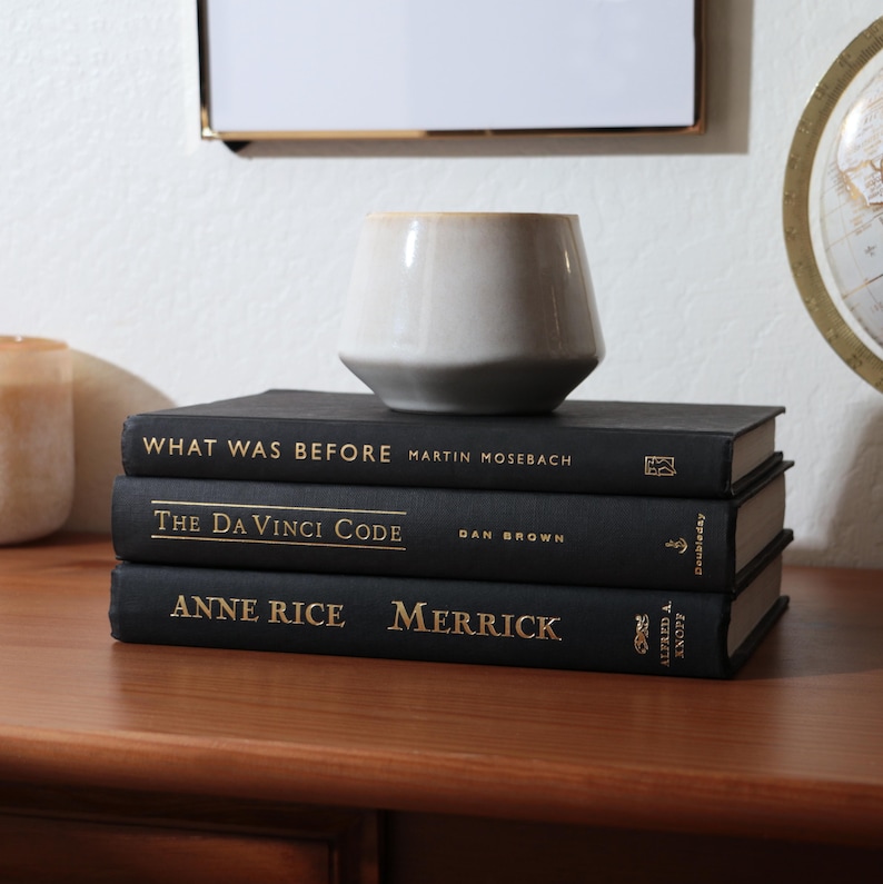 May include: A stack of three black hardcover books with gold lettering, including "What Was Before," "The Da Vinci Code," and "Anne Rice Merrick." A small, light-colored ceramic vase sits atop the books on a wooden surface.