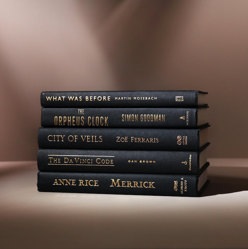 May include: A stack of five hardcover books with black covers and gold lettering. Titles include "What Was Before," "The Orpheus Clock," "City of Veils," "The Da Vinci Code," and "Merrick." The books are arranged on a neutral surface.