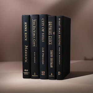 May include: A stack of five hardcover books with black covers and gold lettering. Titles include "Anne Rice Merrick", "The Da Vinci Code", "City of Veils", "The Orpheus Clock", and "What Was Before". The books are arranged vertically.