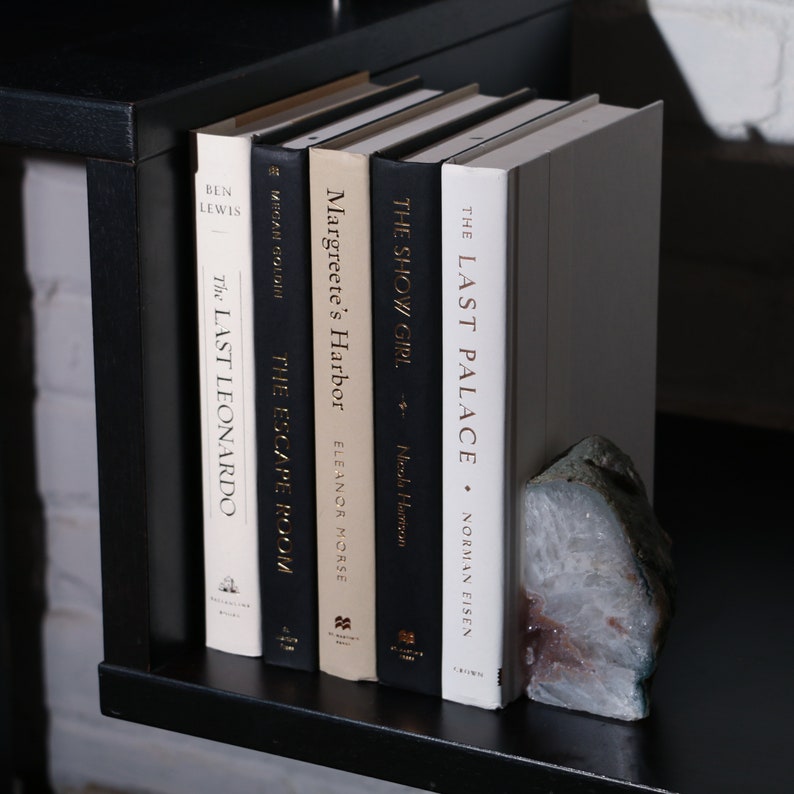 May include: A collection of hardcover books with titles including "The Last Palace" and "The Last Leonardo" on a black shelf. A decorative stone bookend is placed next to the books. The books have various colors and fonts.