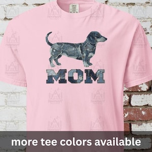 May include: A light pink t-shirt featuring a dachshund dog illustration made of denim patches above the word "MOM" in denim-style letters. The text "more tee colors available" is at the bottom.