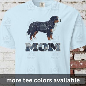 May include: Light blue t-shirt featuring a graphic of a Bernese Mountain Dog with denim-like patches. The word "MOM" is printed below the dog in a denim-style font. The text "more tee colors available" is at the bottom.