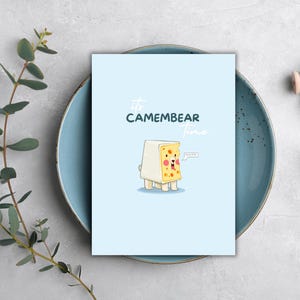 Könnte beinhalten: Eine hellblaue Karte mit dem Text "It's CAMEMBEAR time" und einer Illustration eines Käseblocks mit Bärenmerkmalen. Der Käsebär hat eine Sprechblase. Die Karte liegt auf einem blauen Teller.