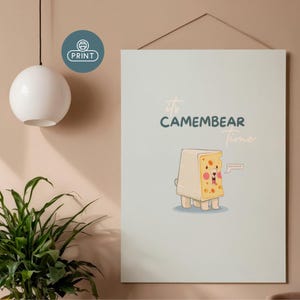 Könnte beinhalten: Ein hellblauer Druck mit einem Cartoon-Camembert-Käse-Charakter mit einem Bärengesicht, dem Text "it's CAMEMBEAR Time" und einer Sprechblase mit der Aufschrift "BONJOUR". Der Druck hängt an einer Wand.