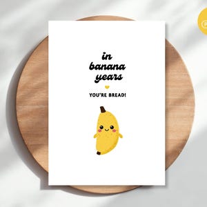 Könnte beinhalten: Eine weiße rechteckige Grußkarte mit dem schwarzen Text in banana years, einem kleinen gelben Herz, dem schwarzen Text YOU'RE BREAD! und einer Cartoon-Illustration einer lächelnden gelben Banane mit rosa Wangen und kleinen Armen.