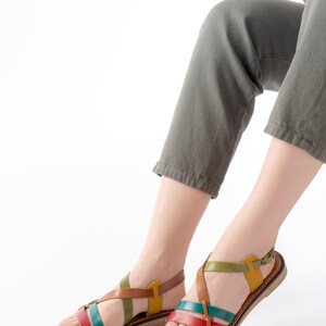 May include: Colorful leather sandals with crisscross straps in red, green, yellow, and brown. The sandals have a flat sole and a buckle closure. The image shows the sandals on a person's feet, with olive green pants and a white background.