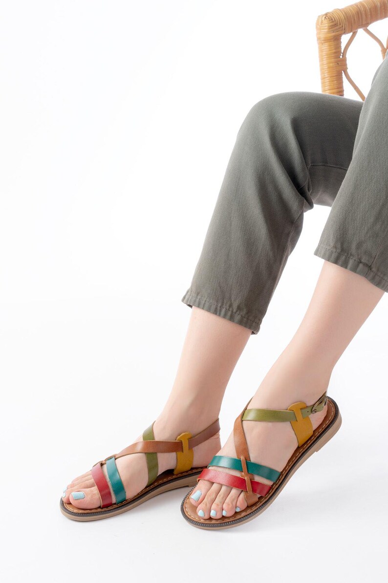 May include: Colorful leather sandals with crisscross straps in red, green, yellow, and brown. The sandals have a flat sole and a buckle closure. The image shows the sandals on a person's feet, with olive green pants and a white background.