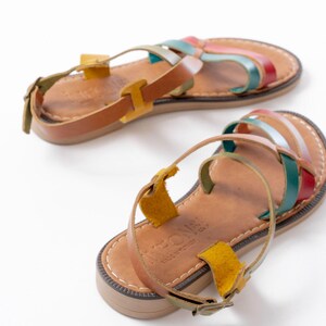 May include: A pair of leather sandals with colorful straps in red, green, blue, and pink. The sandals have a brown footbed and a buckle closure. The sandals are displayed on a white background.