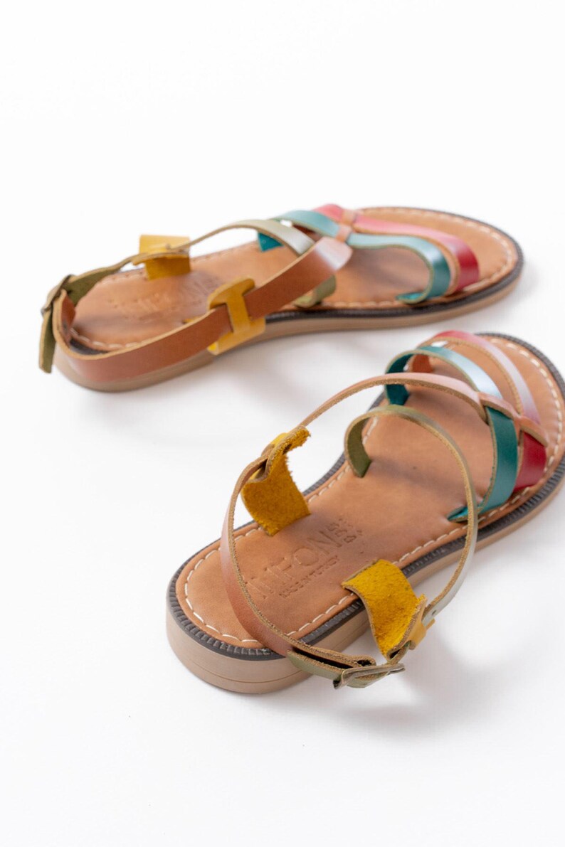 May include: A pair of leather sandals with colorful straps in red, green, blue, and pink. The sandals have a brown footbed and a buckle closure. The sandals are displayed on a white background.
