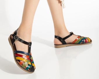 May include: Colorful leather sandals with a woven design. The sandals feature a black ankle strap and a multi-colored woven pattern in red, yellow, green, and blue. The soles are a light brown color.