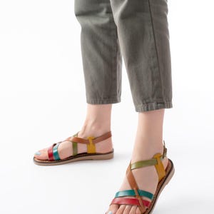 May include: Leather sandals with colorful straps in red, teal, green, and yellow. The sandals have a brown sole and a buckle closure. The person is wearing olive green pants.