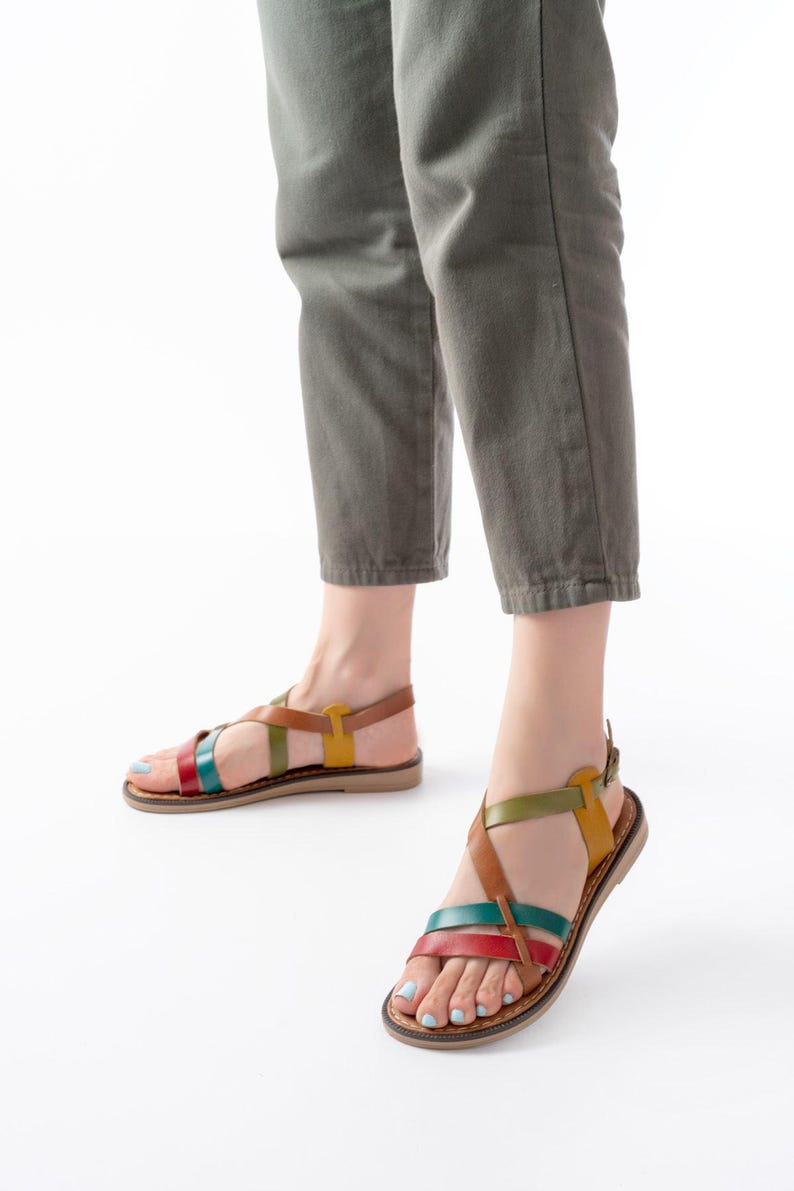 May include: Leather sandals with colorful straps in red, teal, green, and yellow. The sandals have a brown sole and a buckle closure. The person is wearing olive green pants.