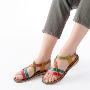May include: Multi-colored leather sandals with a brown sole. The sandals feature straps in red, green, brown, and yellow. The ankle strap has a buckle closure. The sandals are shown on a white background.