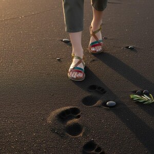 May include: A person wearing brown, red, and teal leather sandals with gold buckles walks on dark sand, leaving footprints. The person is wearing olive green pants. The scene is lit by warm sunlight.
