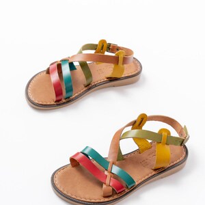 May include: Two pairs of leather sandals with colorful straps. The sandals have a brown sole and a strap that buckles around the ankle. The straps are red, teal, green, brown, and yellow. The sandals are displayed on a white background.