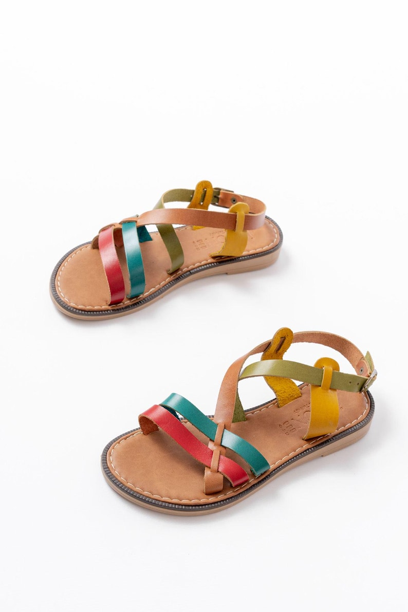May include: Two pairs of leather sandals with colorful straps. The sandals have a brown sole and a strap that buckles around the ankle. The straps are red, teal, green, brown, and yellow. The sandals are displayed on a white background.