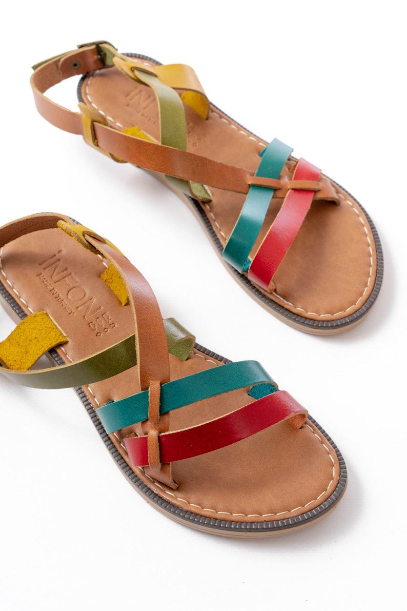 May include: Leather sandals with a multi-colored strap design. The sandals feature straps in yellow, green, red, and teal. The footbed is a light brown color with a stitched border. The sandals have a buckle closure and the text "INFONE" is visible.