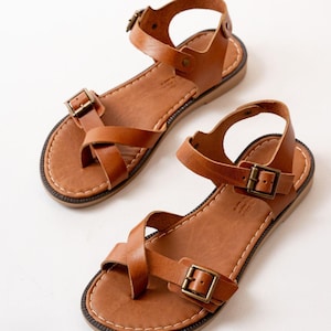 May include: A pair of tan leather sandals featuring a toe loop design, an adjustable ankle strap with a metal buckle, and a secondary strap across the forefoot with a matching metal buckle. The footbed is stitched with white thread along the edge.