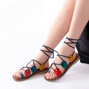 May include: Colorful leather sandals with a wrap-around ankle design. The sandals feature red, blue, green, and yellow heart-shaped accents and a brown sole. The navy blue laces wrap around the ankles and tie at the top.